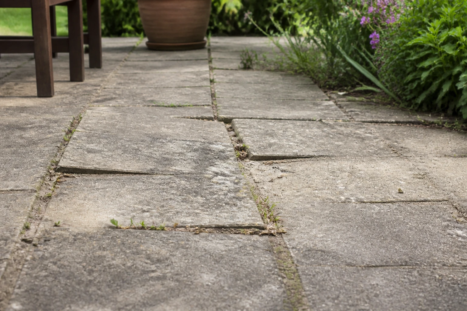 Dalles de terrasse posées sur sable avec joints érodés, surface irrégulière et mauvaises herbes entre les pierres